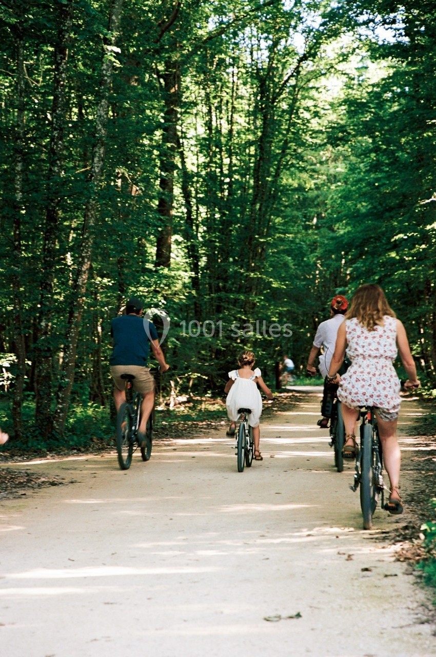 Des personnes, dont une enfant, font du vélo sur un chemin forestier ombragé par une journée ensoleillée.