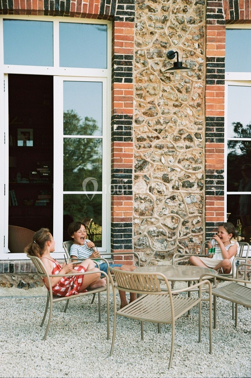 Trois enfants assis autour d'une table en extérieur, devant un mur en pierre et des fenêtres, discutant et riant.