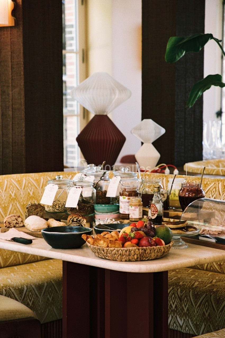 Table garnie de divers aliments pour un buffet, incluant fruits, confitures, pains et céréales, dans un cadre chaleureux.