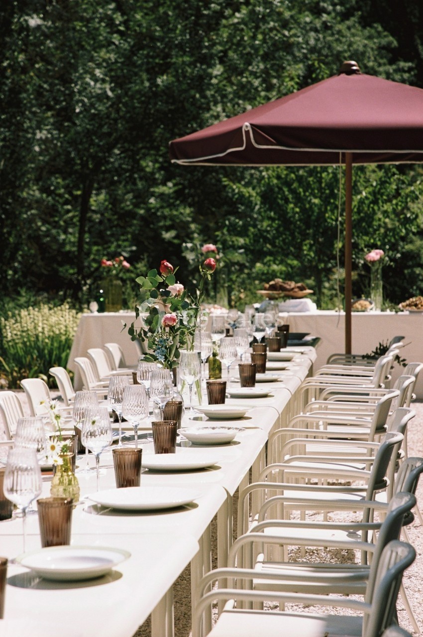 Grande table extérieure dressée avec vaisselle et décorations florales, sous un parasol rouge, dans un jardin verdoyant.