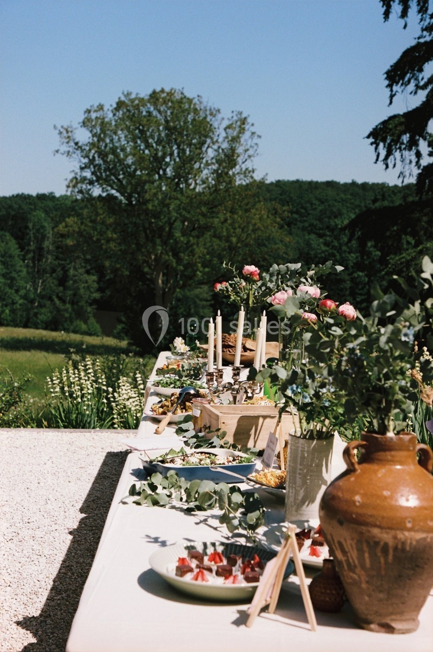 Table dressée en extérieur avec plats variés, bougies et décorations florales, entourée de verdure.