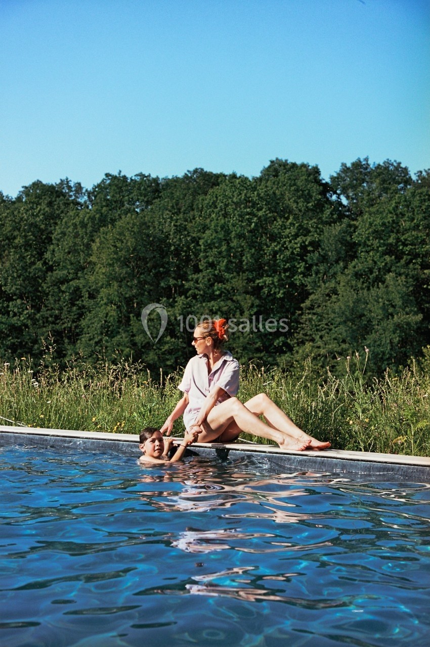 Une femme assise au bord d'une piscine aide un enfant dans l'eau, avec une forêt en arrière-plan.