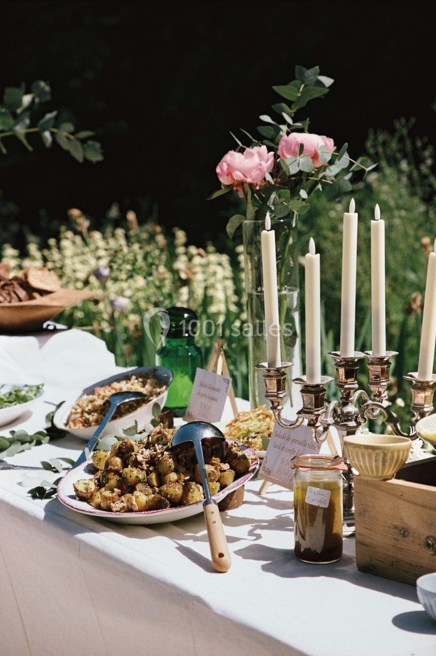 Buffet en plein air avec plats variés, bougies, fleurs et condiments sur une table blanche dans un cadre verdoyant.