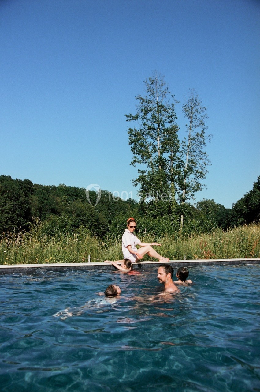 Des personnes se baignent dans une piscine extérieure entourée de verdure sous un ciel bleu.