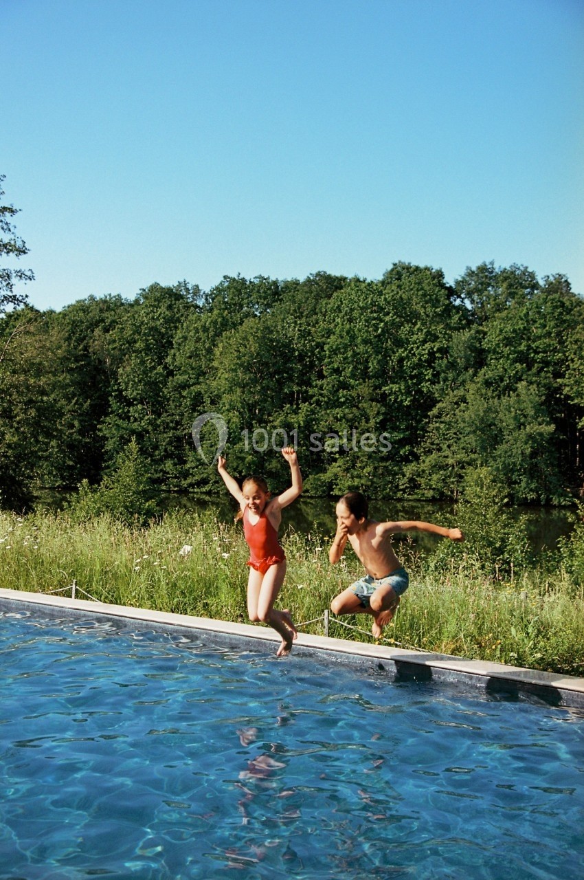 Deux personnes sautent dans une piscine en plein air, entourée de verdure et d'arbres sous un ciel dégagé.