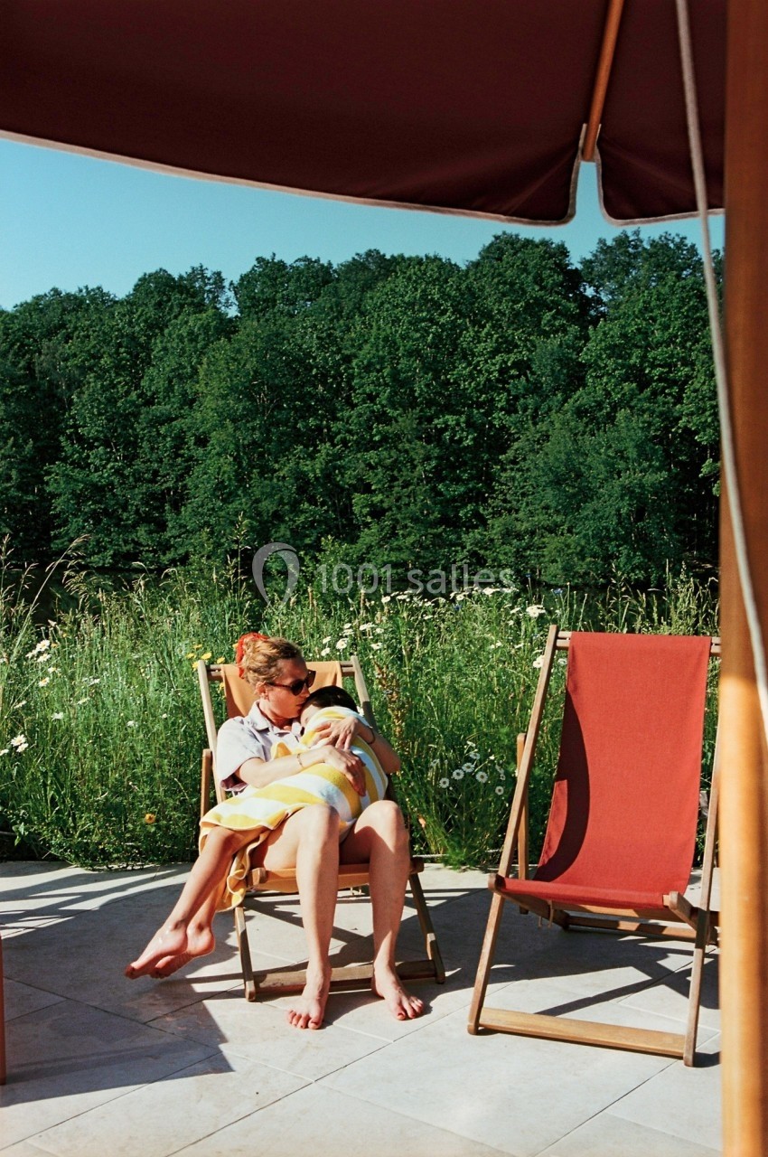 Une femme assise sur une chaise longue tient un bébé dans ses bras, entourée de verdure et de chaises vides.