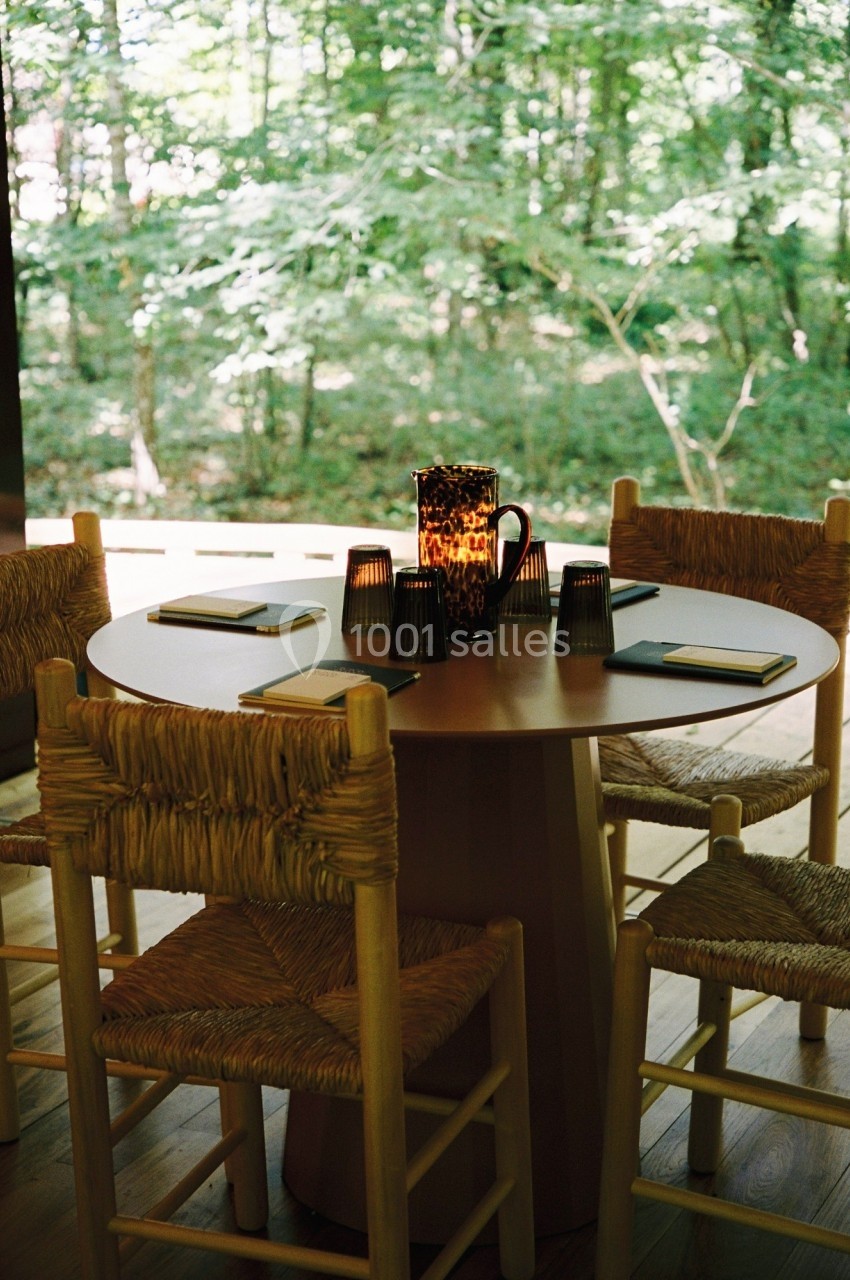 Table ronde en bois entourée de chaises en paille, avec des carnets et une carafe au centre, vue sur une forêt.