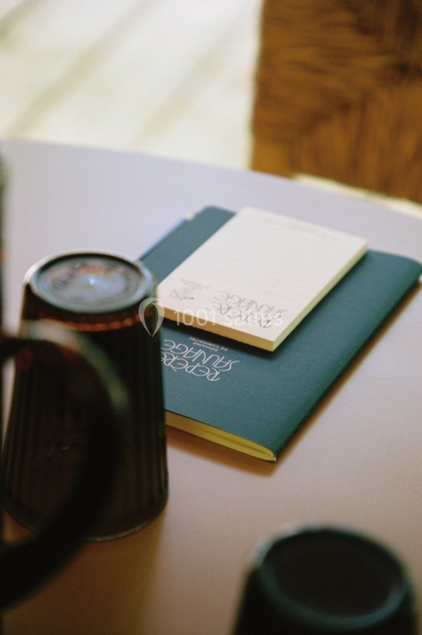 Carnet bleu et bloc-notes posés sur une table, entourés de tasses floues au premier plan.