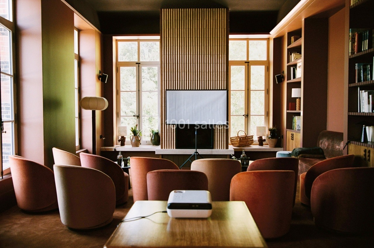 Salle de projection lumineuse avec fauteuils colorés, écran blanc, projecteur et étagères remplies de livres.