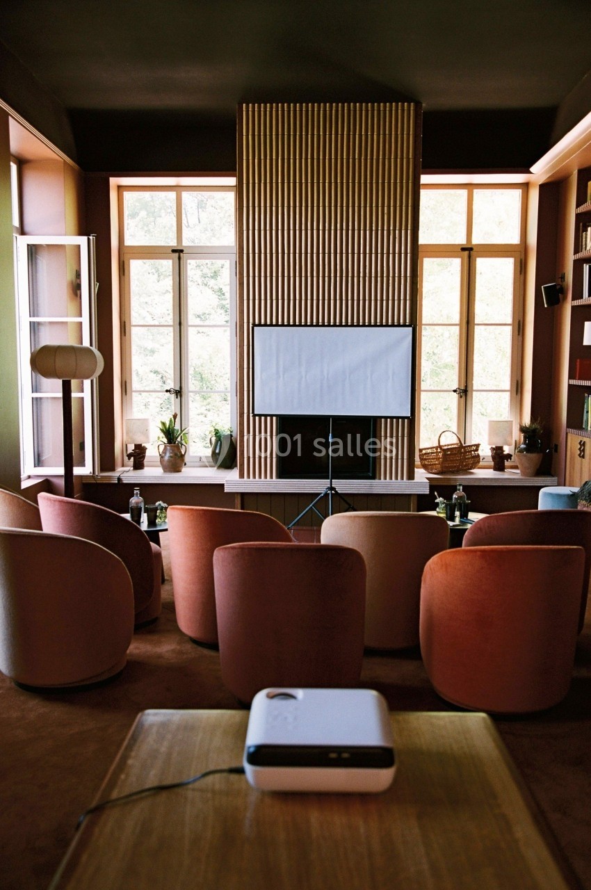 Salle aménagée avec des fauteuils colorés, un projecteur et un écran blanc devant de grandes fenêtres.