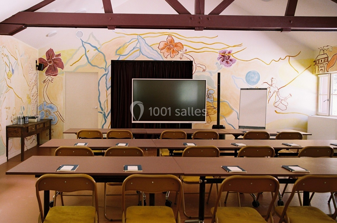 Salle de réunion avec tables, chaises alignées, écran central, tableau blanc et fresques murales colorées.