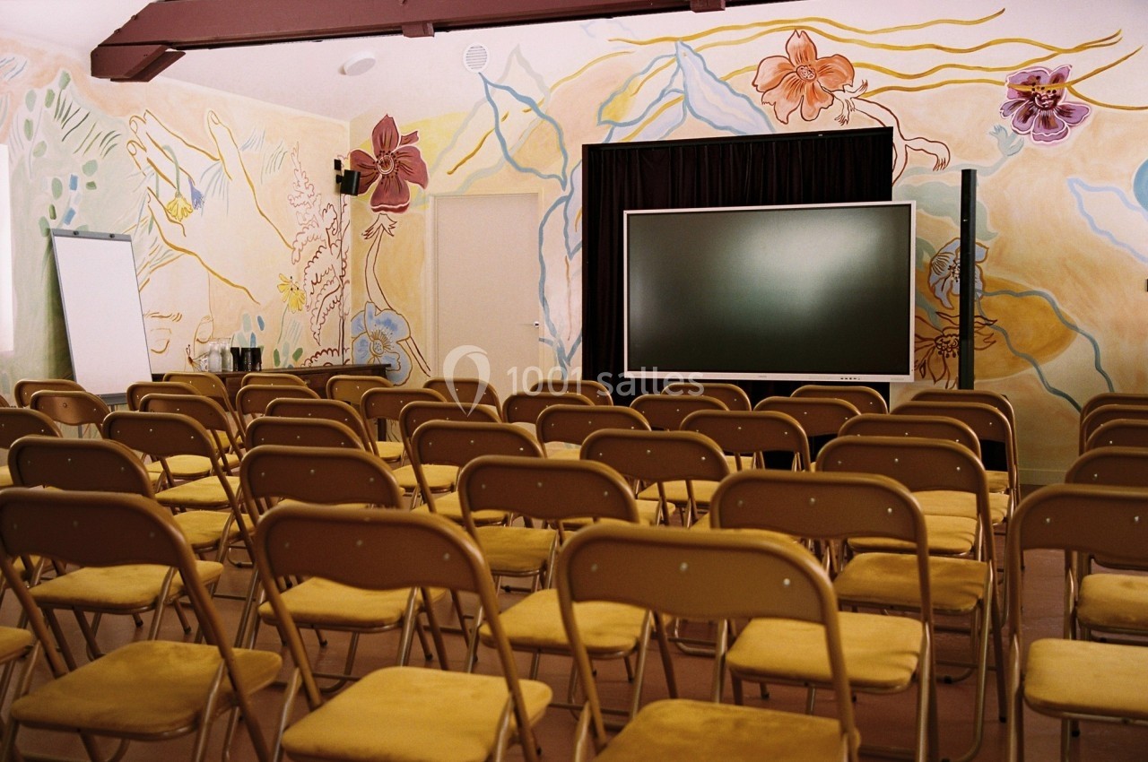 Salle de conférence avec des rangées de chaises pliantes jaunes, un écran noir et des murs décorés de peintures colorées.