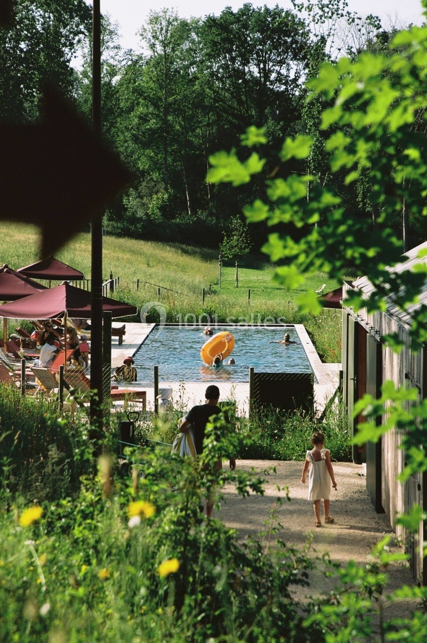 Vue d'une piscine extérieure entourée de verdure, avec des personnes se relaxant et des enfants jouant.