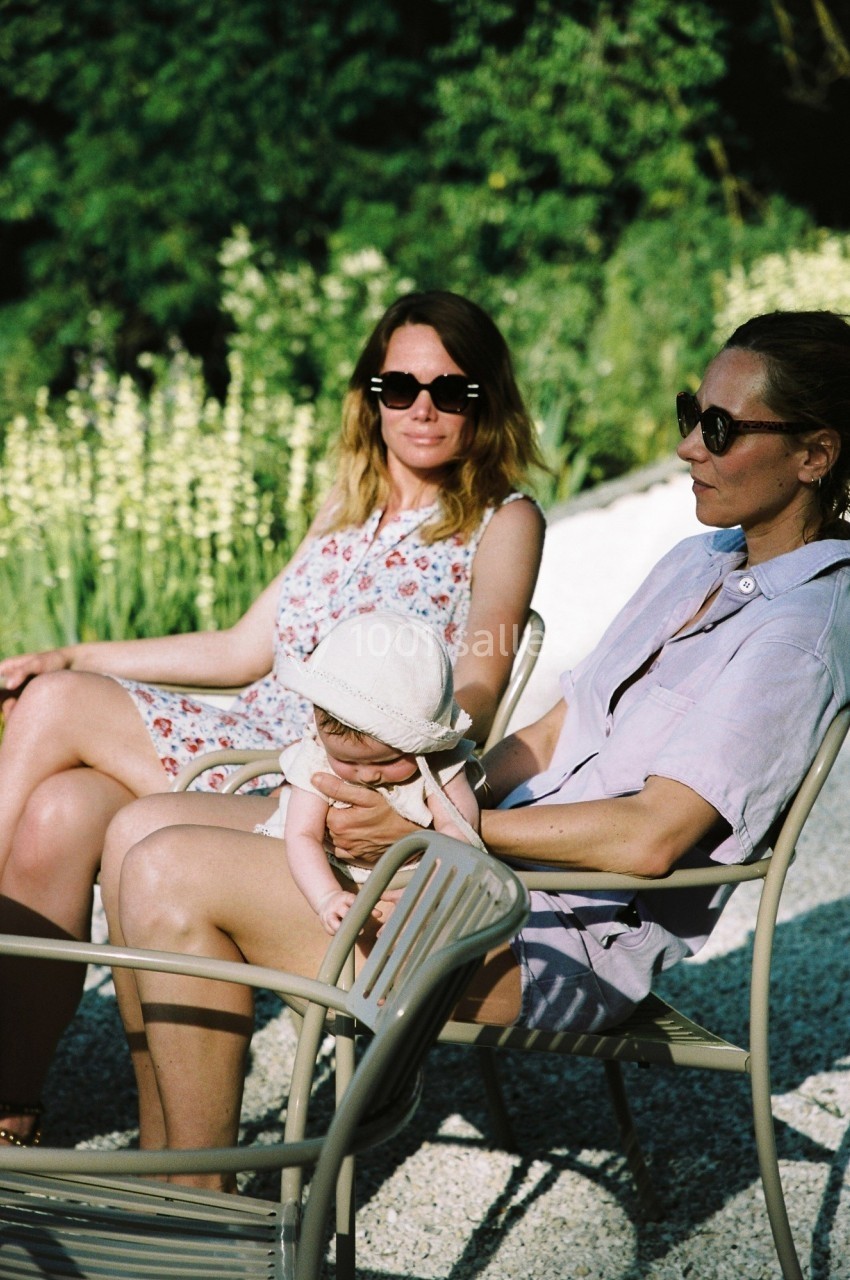Deux femmes assises sur des chaises de jardin, l'une tenant un bébé portant un chapeau, avec un fond de verdure.
