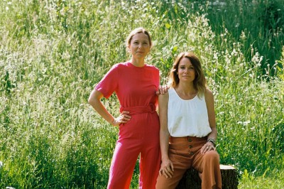 Deux femmes posent dans un jardin verdoyant devant un bâtiment en briques avec une porte et un escalier en colimaçon.