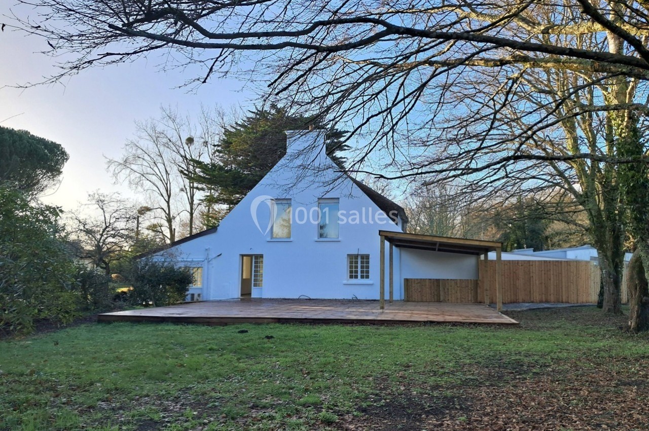Maison blanche avec toit en pente, terrasse en bois et jardin arboré, sous un ciel dégagé en fin de journée.