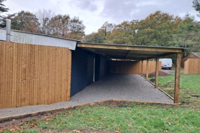 Abri en bois avec toit en pente, sol en gravier et clôture attenante, entouré de végétation et d'herbe.