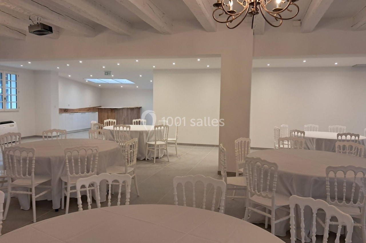 Salle lumineuse avec tables rondes dressées de nappes blanches, chaises blanches et un lustre au plafond.