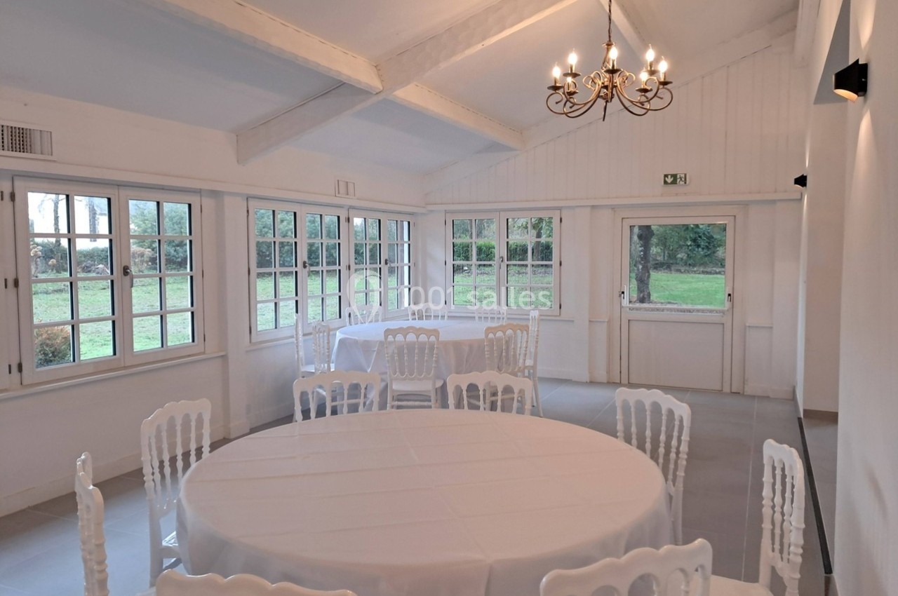 Salle lumineuse avec tables rondes recouvertes de nappes blanches, chaises blanches et vue sur un jardin à travers de…