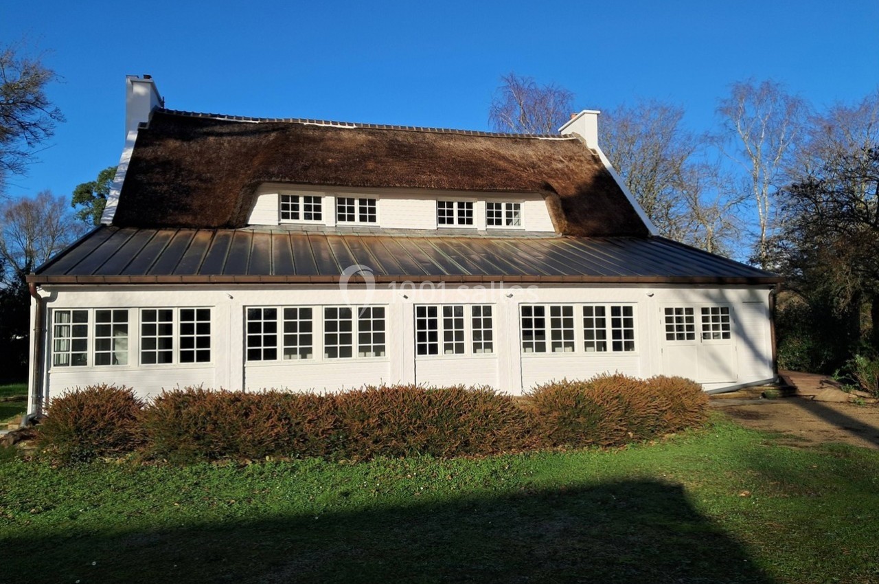 Maison blanche avec toit de chaume, grandes fenêtres et jardin, entourée d'arbres sous un ciel bleu.