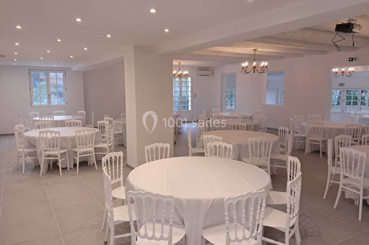 Salle lumineuse avec tables rondes dressées de nappes blanches et chaises blanches, éclairée par des lustres et des spots.