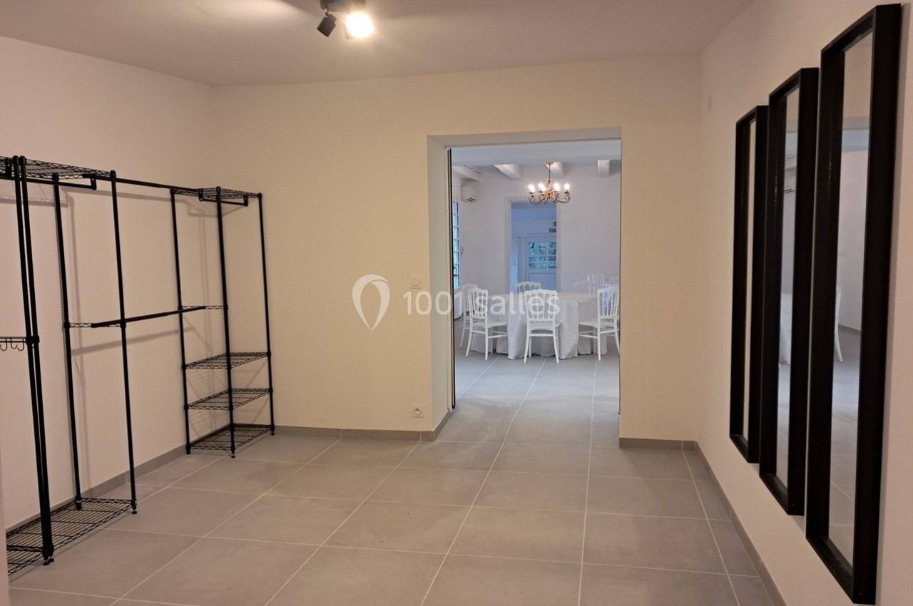 Pièce vide avec étagères métalliques à gauche, miroirs à droite et vue sur une salle avec chaises blanches au fond.