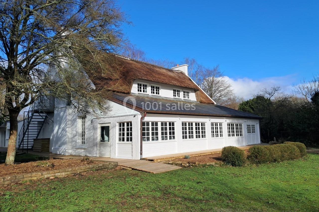 Maison blanche avec toit de chaume, entourée de verdure, sous un ciel bleu dégagé.
