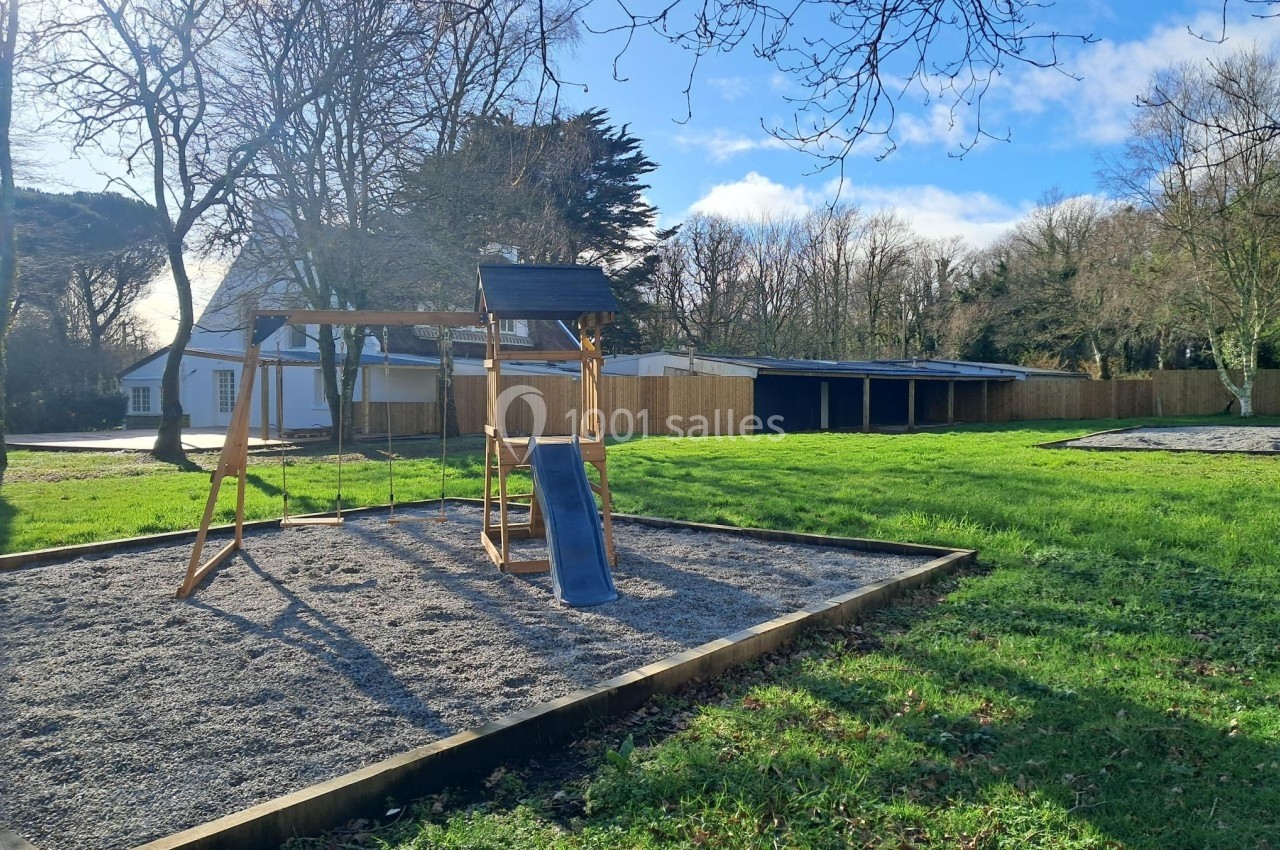Aire de jeux avec balançoire et toboggan sur sol en gravier, entourée de pelouse et d'arbres sous un ciel dégagé.