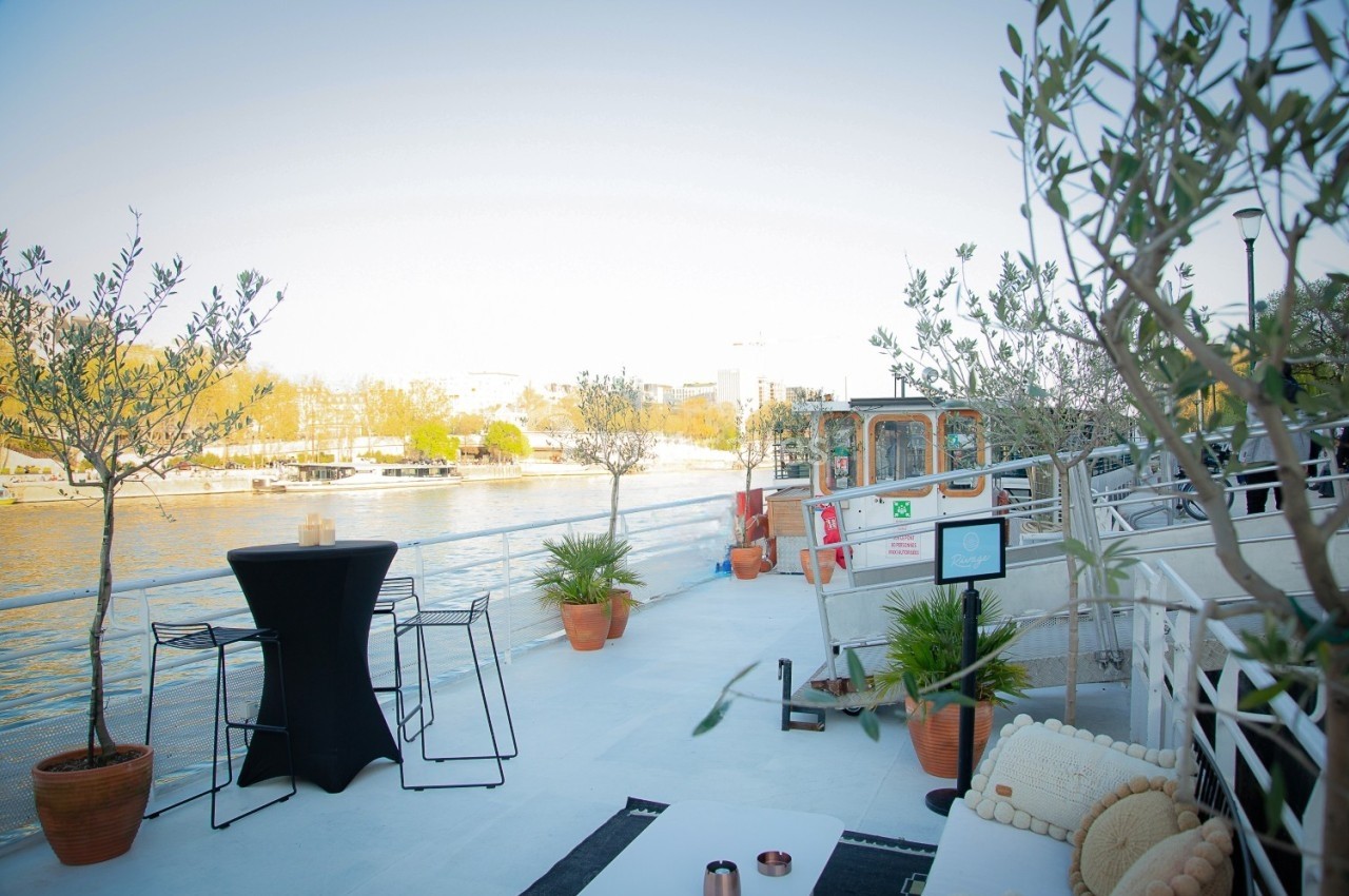 Terrasse extérieure d'une péniche aménagée avec des plantes en pots, des tables hautes et des chaises, au bord d'une rivière.