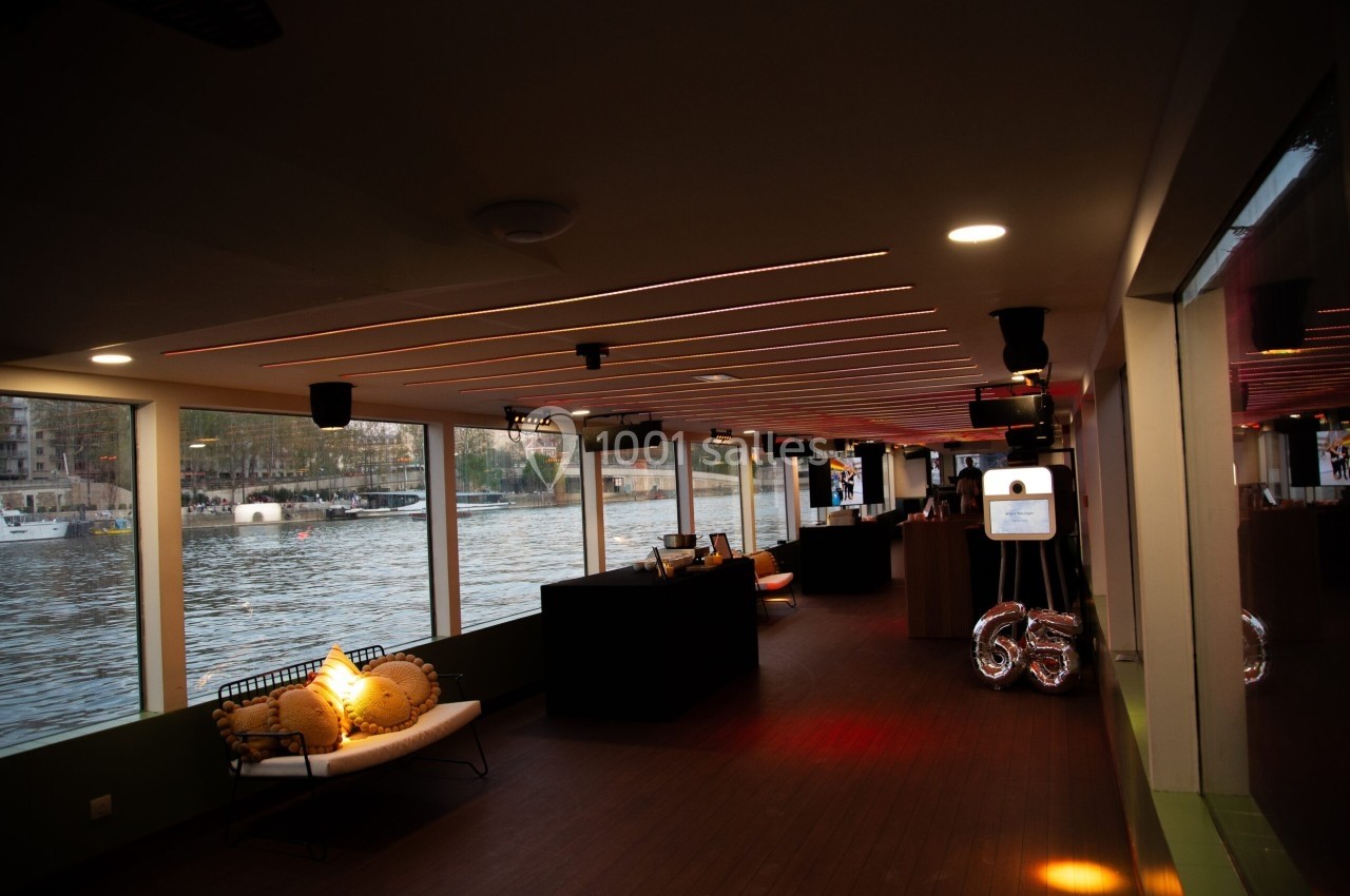 Intérieur d'une péniche avec vue sur l'eau, éclairage tamisé et mobilier moderne le long des baies vitrées.