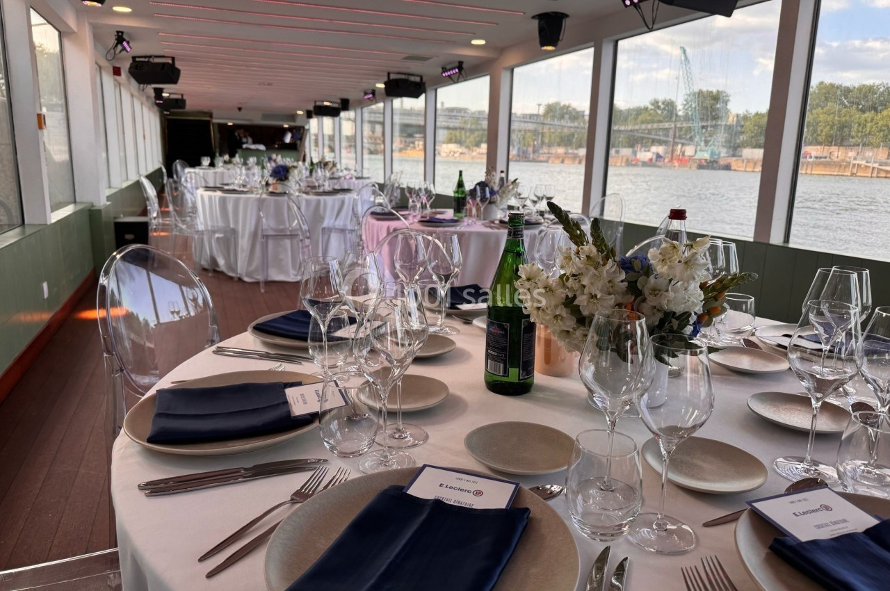 Salle de réception élégante avec tables dressées, nappes blanches, serviettes bleues et vue sur un fleuve à travers de…