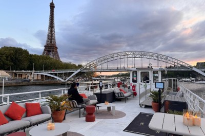 Miniature Location salle Paris 5 (Paris) - Péniche Amour #8 Terrasse d'un bateau aménagée avec des tables et des sièges, vue sur la tour Eiffel et un pont au coucher du soleil.