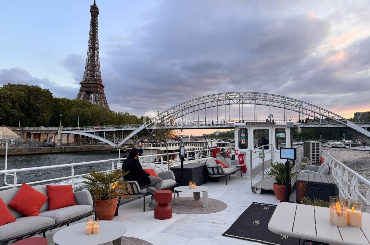 Terrasse d'un bateau aménagée avec des tables et des sièges, vue sur la tour Eiffel et un pont au coucher du soleil.