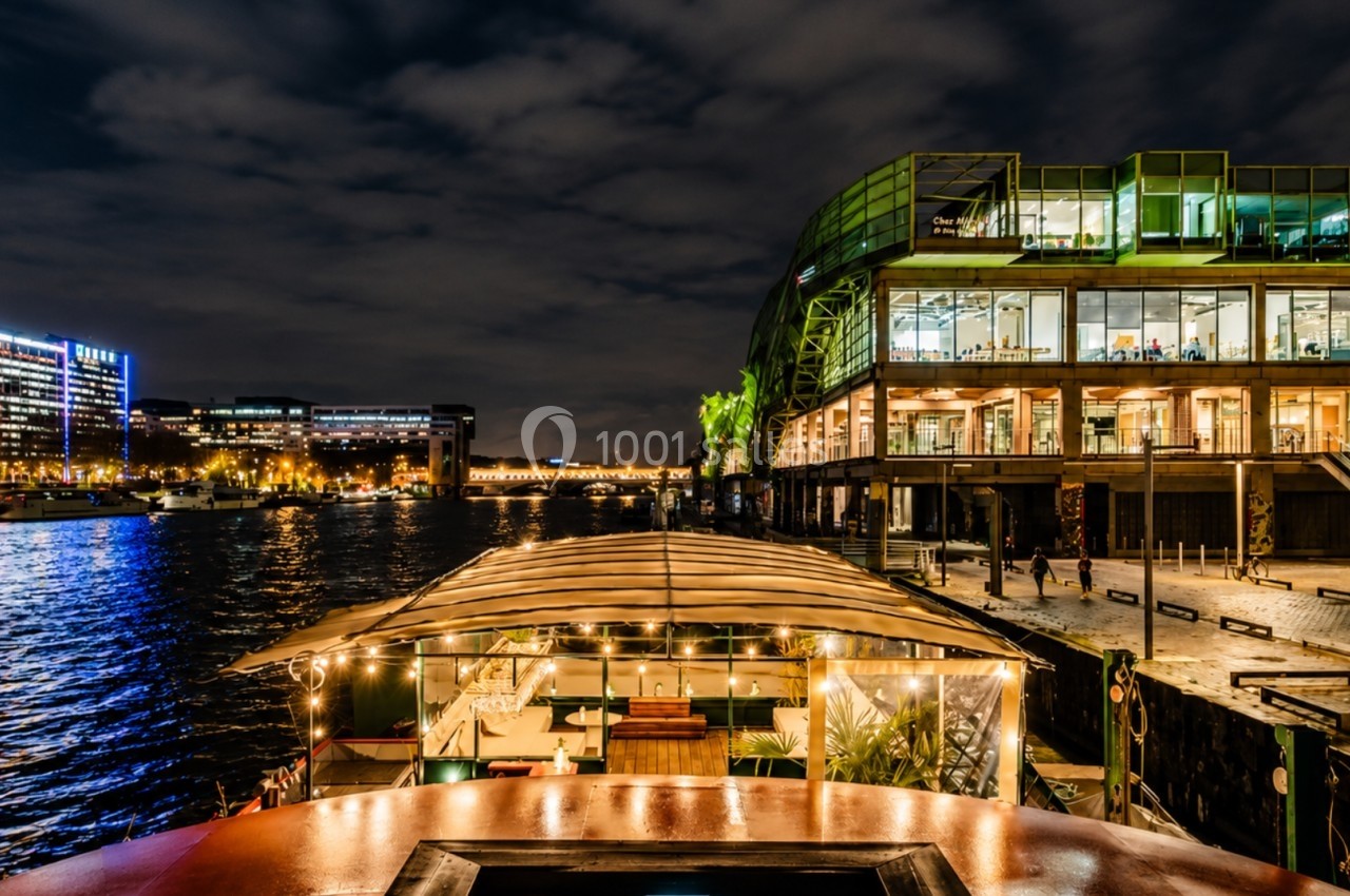 Vue nocturne d'un quai animé avec un bâtiment moderne éclairé et une péniche illuminée sur une rivière calme.
