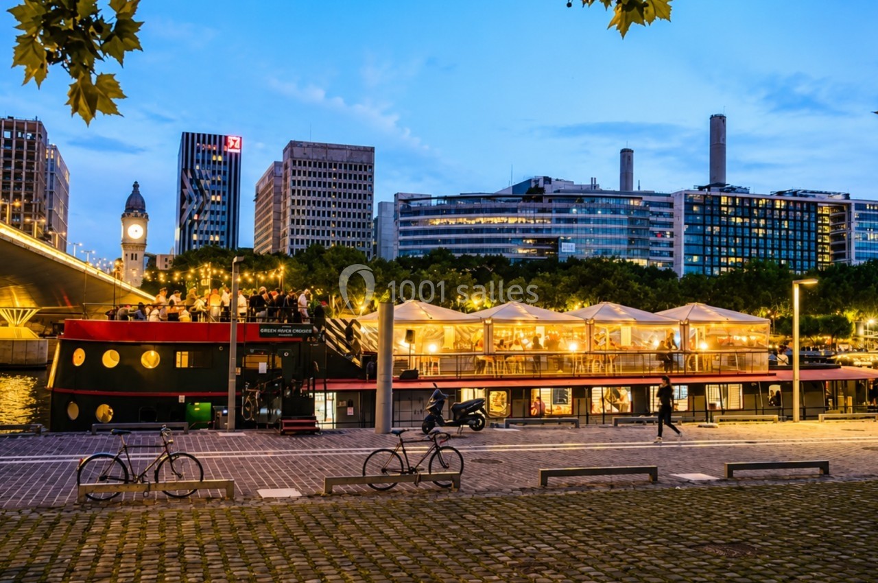 Bateau-restaurant illuminé amarré le long d'un quai au crépuscule, avec des immeubles en arrière-plan.