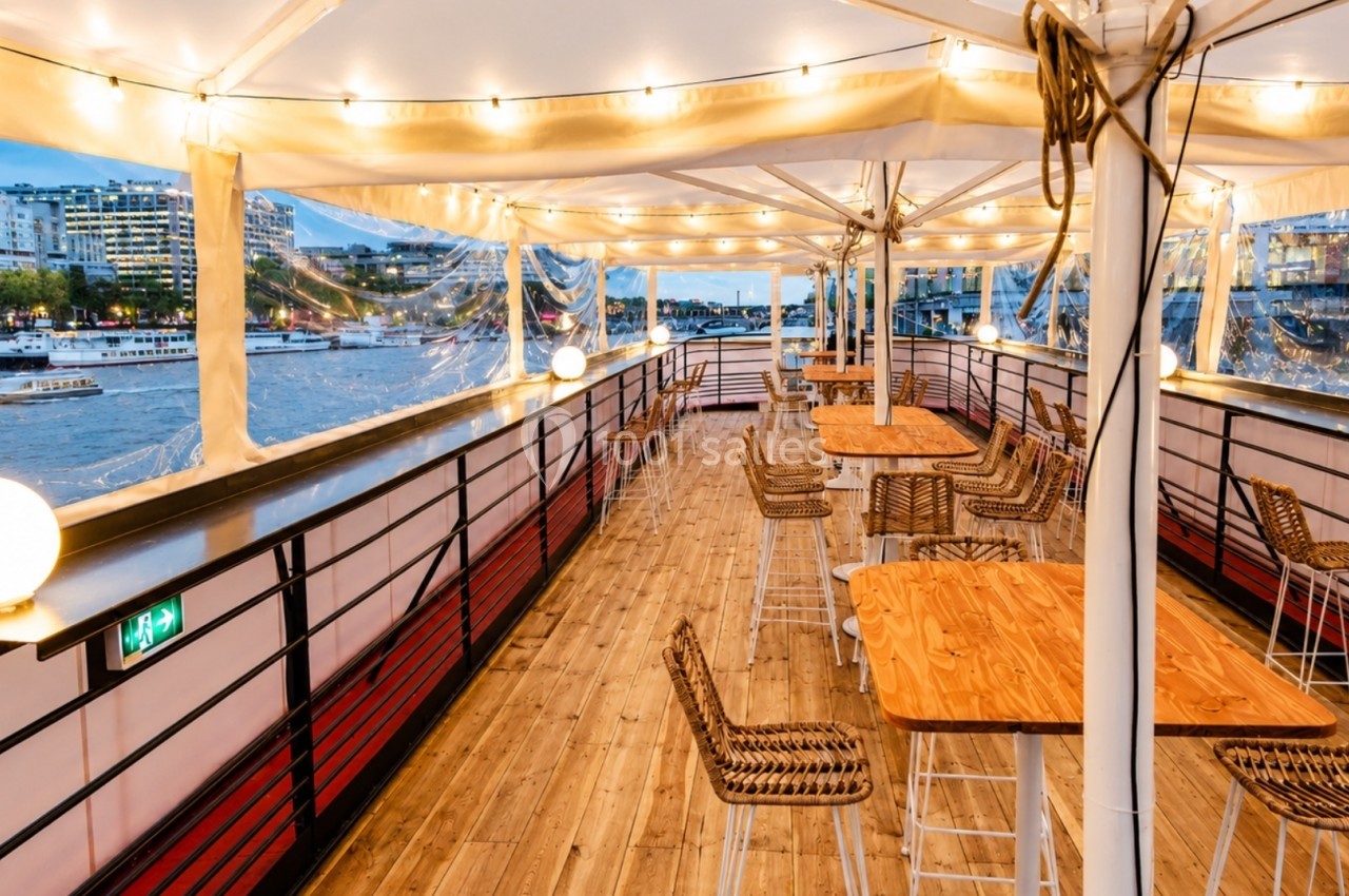 Terrasse couverte d'un bateau avec des tables en bois, des chaises en rotin et une vue sur un fleuve bordé de bâtiments.