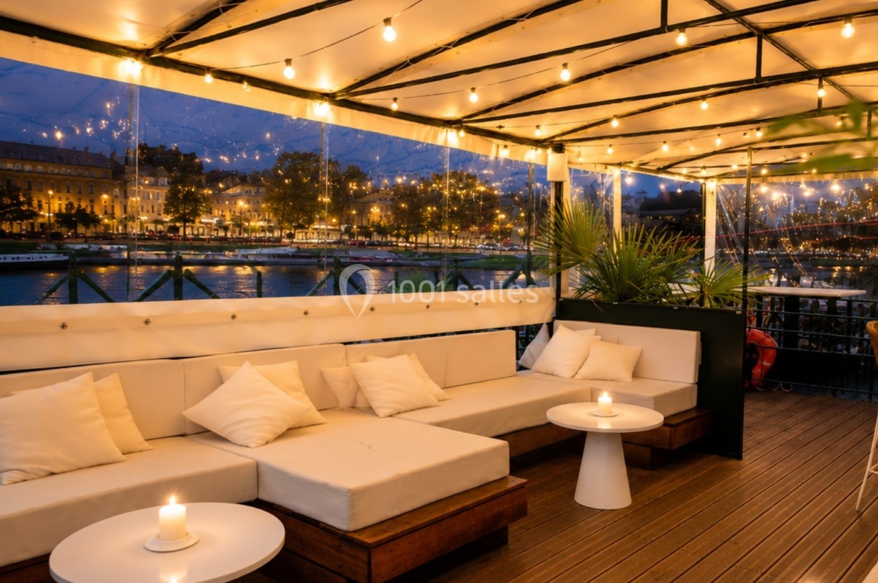 Terrasse cosy en soirée avec canapés, tables basses, bougies allumées et vue sur une rivière illuminée.