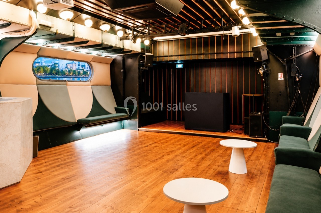 Salle moderne avec parquet, banquettes vertes, tables basses blanches et hublot offrant une vue extérieure.