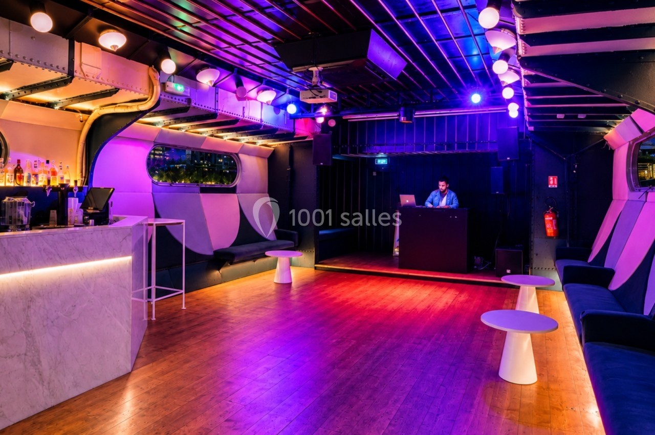 Salle de soirée moderne avec bar, piste de danse, DJ en cabine et éclairage coloré.