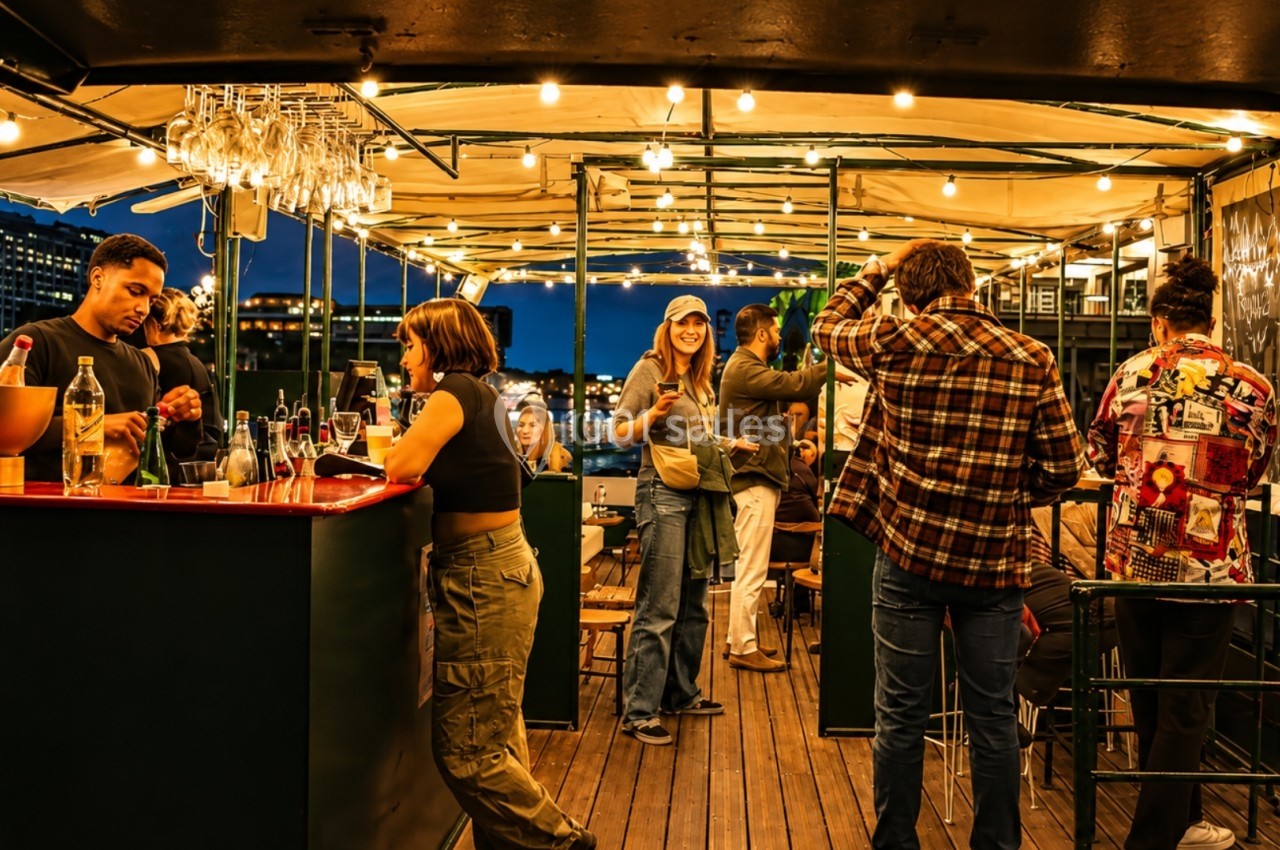 Personnes discutant et partageant des boissons sur une terrasse éclairée en soirée, avec un bar en arrière-plan.