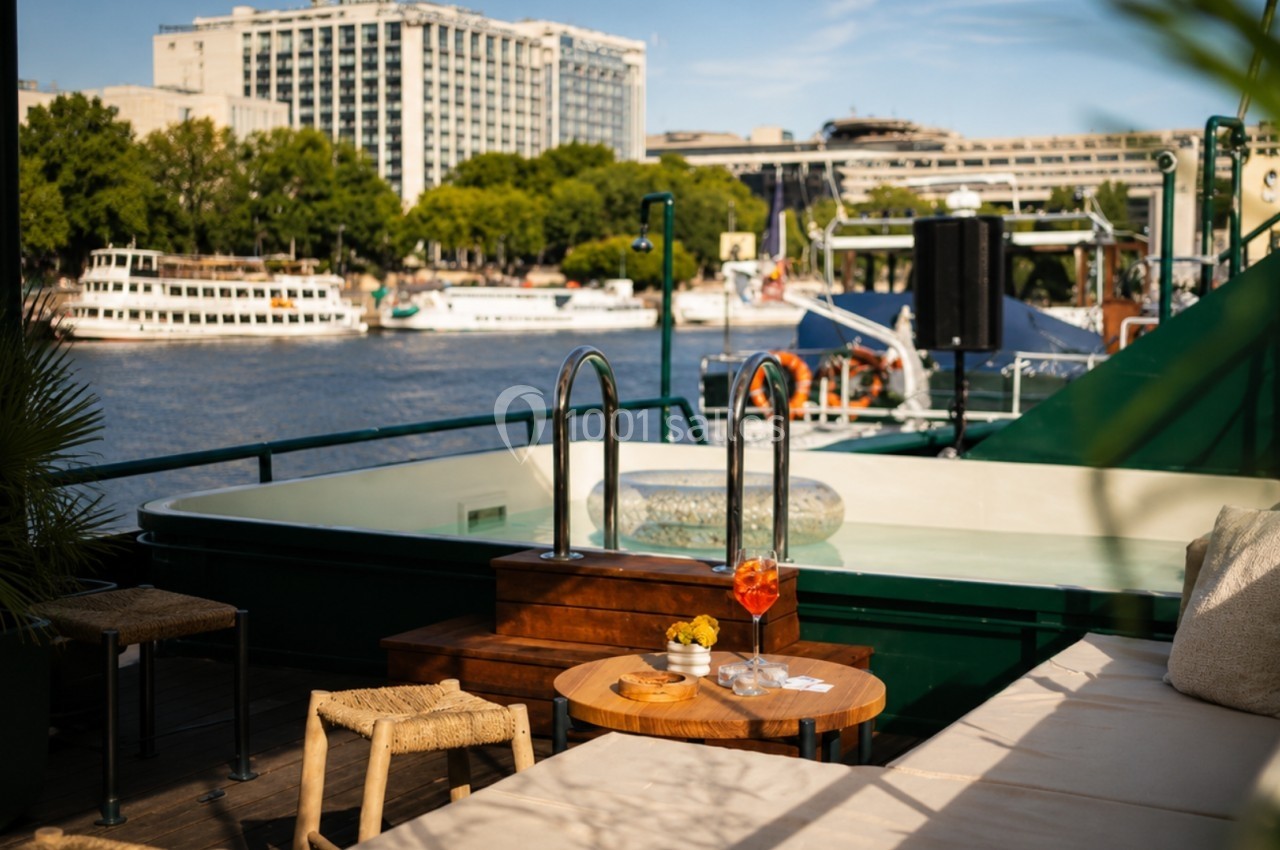 Terrasse avec piscine, table basse et fauteuils, vue sur une rivière bordée de bateaux et bâtiments urbains.