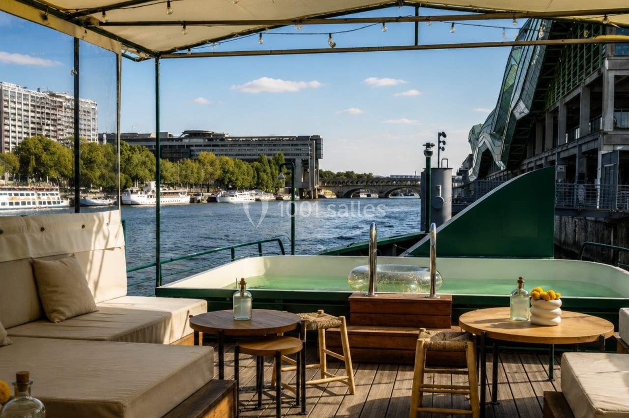 Terrasse aménagée sur une péniche avec vue sur la Seine, des bâtiments et un pont en arrière-plan.