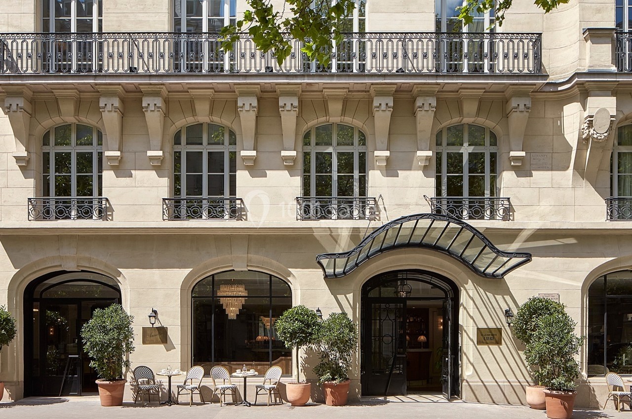 Façade d'un bâtiment haussmannien avec des tables en terrasse et des plantes en pots devant les fenêtres.