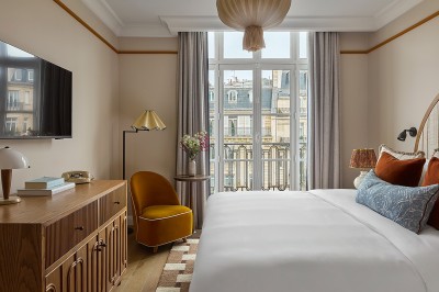 Chambre lumineuse avec lit double, fauteuil jaune, commode en bois et grande fenêtre donnant sur des immeubles parisiens.