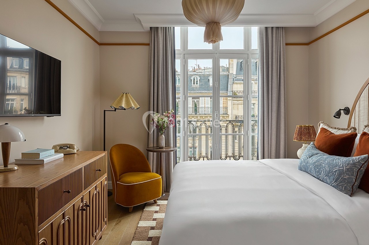 Chambre lumineuse avec lit double, fauteuil jaune, commode en bois et grande fenêtre donnant sur des immeubles parisiens.