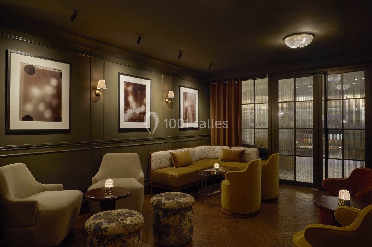 Salon cosy avec fauteuils, banquette, tables basses, éclairage tamisé et tableaux accrochés aux murs sombres.