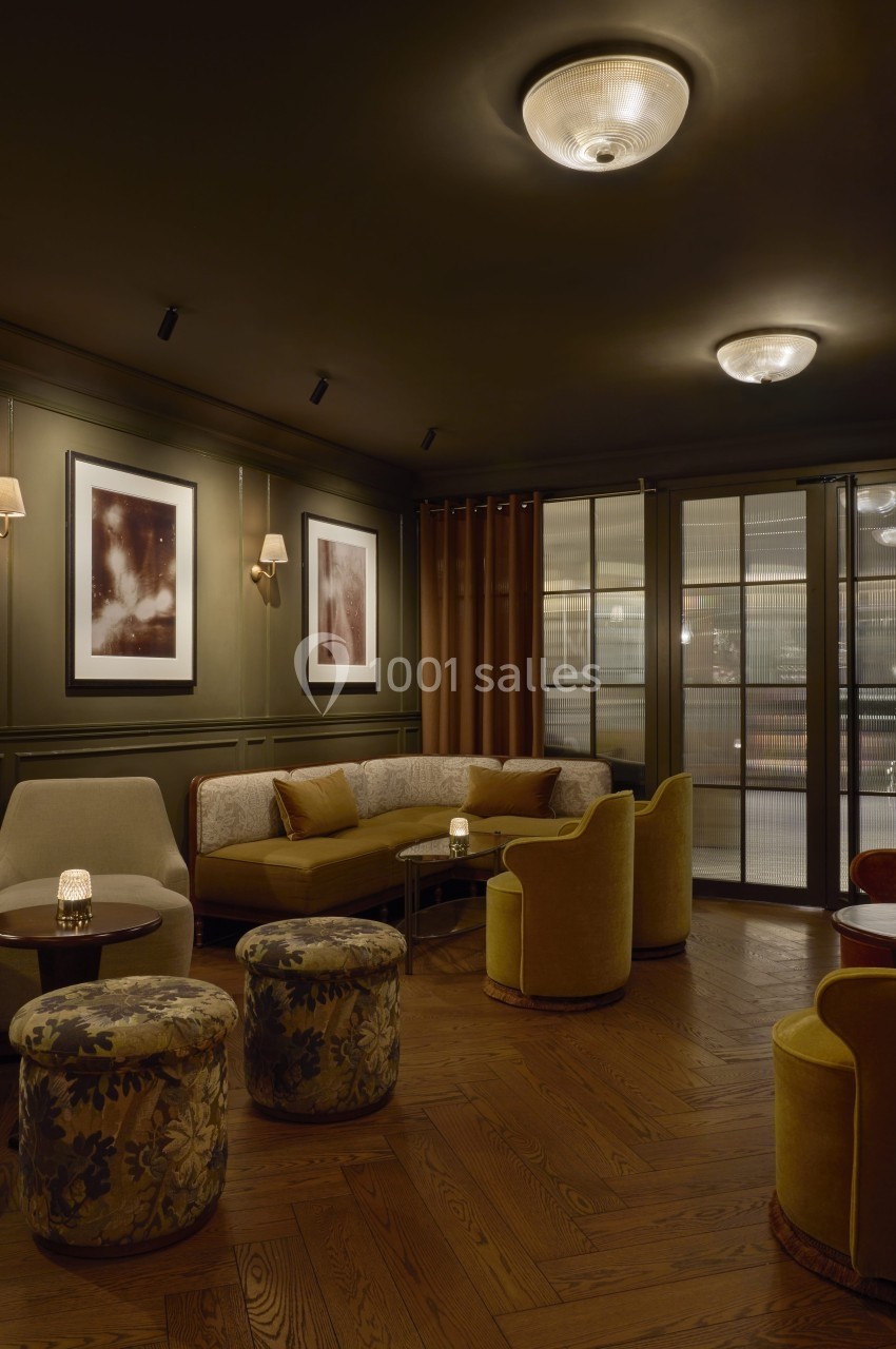 Salon élégant avec fauteuils et poufs colorés, éclairage tamisé, murs verts ornés de tableaux et sol en bois.