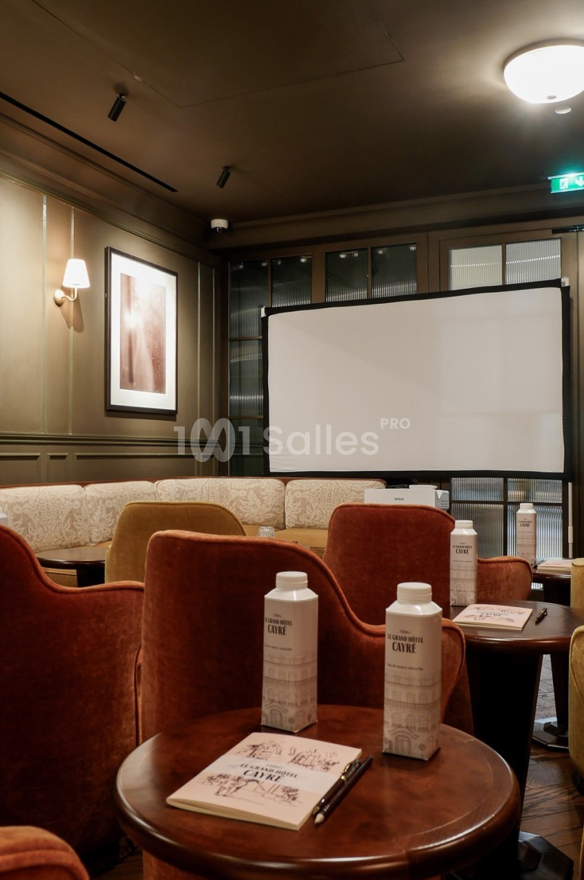Salle de réunion confortable avec fauteuils en velours, tables en bois, écran de projection et carnets disposés.