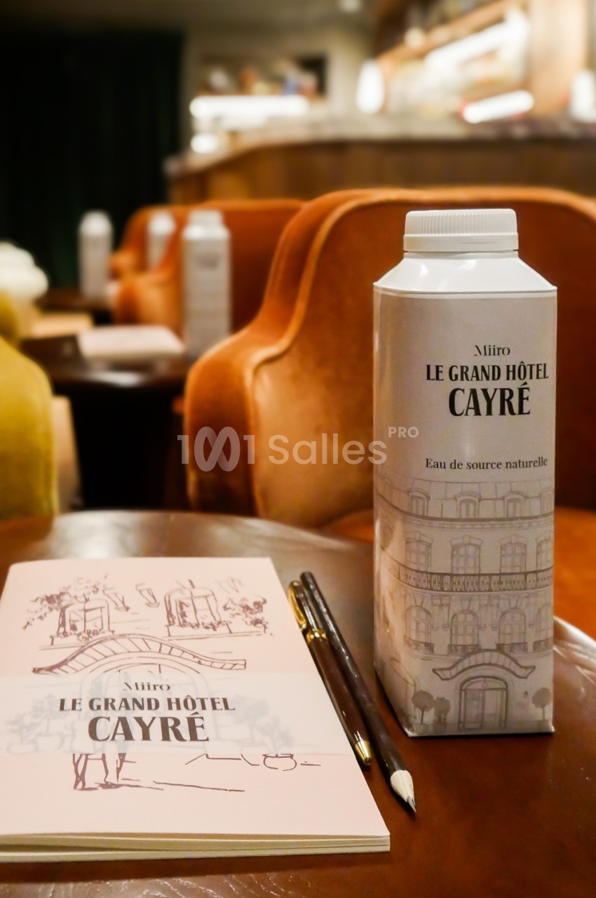 Carton d'eau marqué ’Le Grand Hôtel Cayré’ posé sur une table avec un carnet et des crayons, dans un salon élégant.
