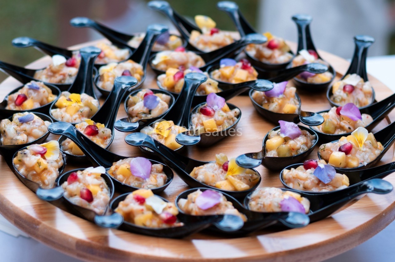 Cuillères noires contenant des portions de tartare de poisson décorées de fleurs comestibles et de morceaux de fruits.